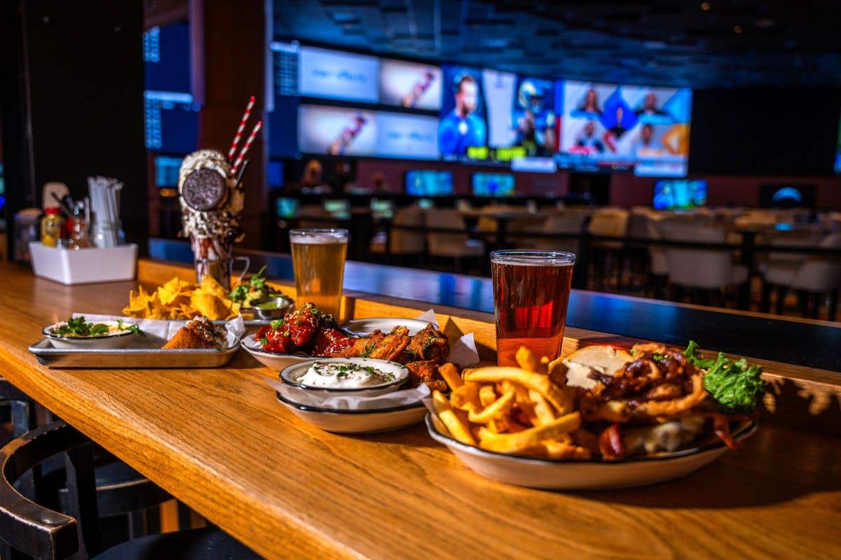 Burger, fries, wings, and beer on a bar with multiple game screens in the background