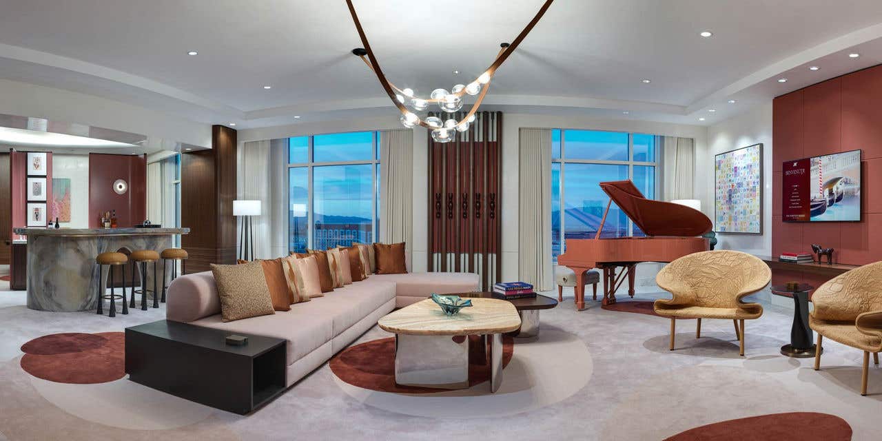 A large hotel living room at night with a couch, coffee table, baby grand piano, chandelier, and bar seating.